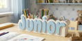 A wooden desk organizer shaped like the name 'Junior' in light blue letters on a white base, filled with colored pencils and small wooden toys, set in a child's room with books and stuffed animals in the background.